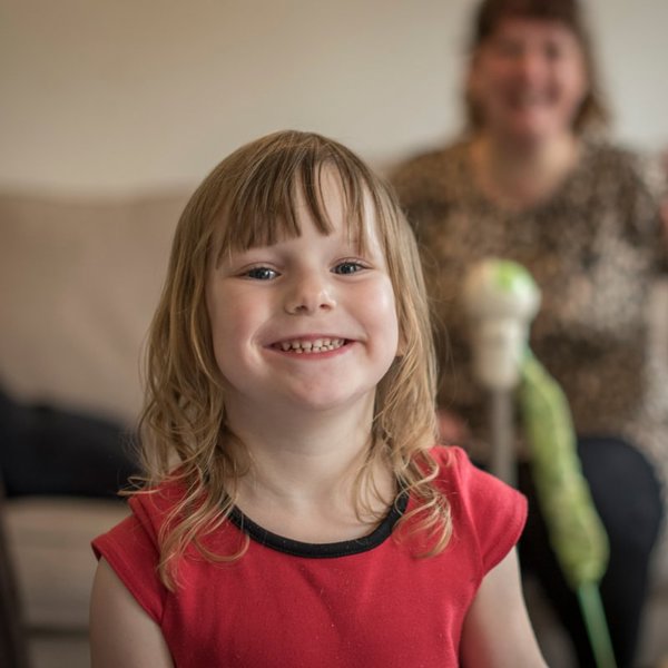 A young child in a red teeshirt smiling broadly from the inside of her home
