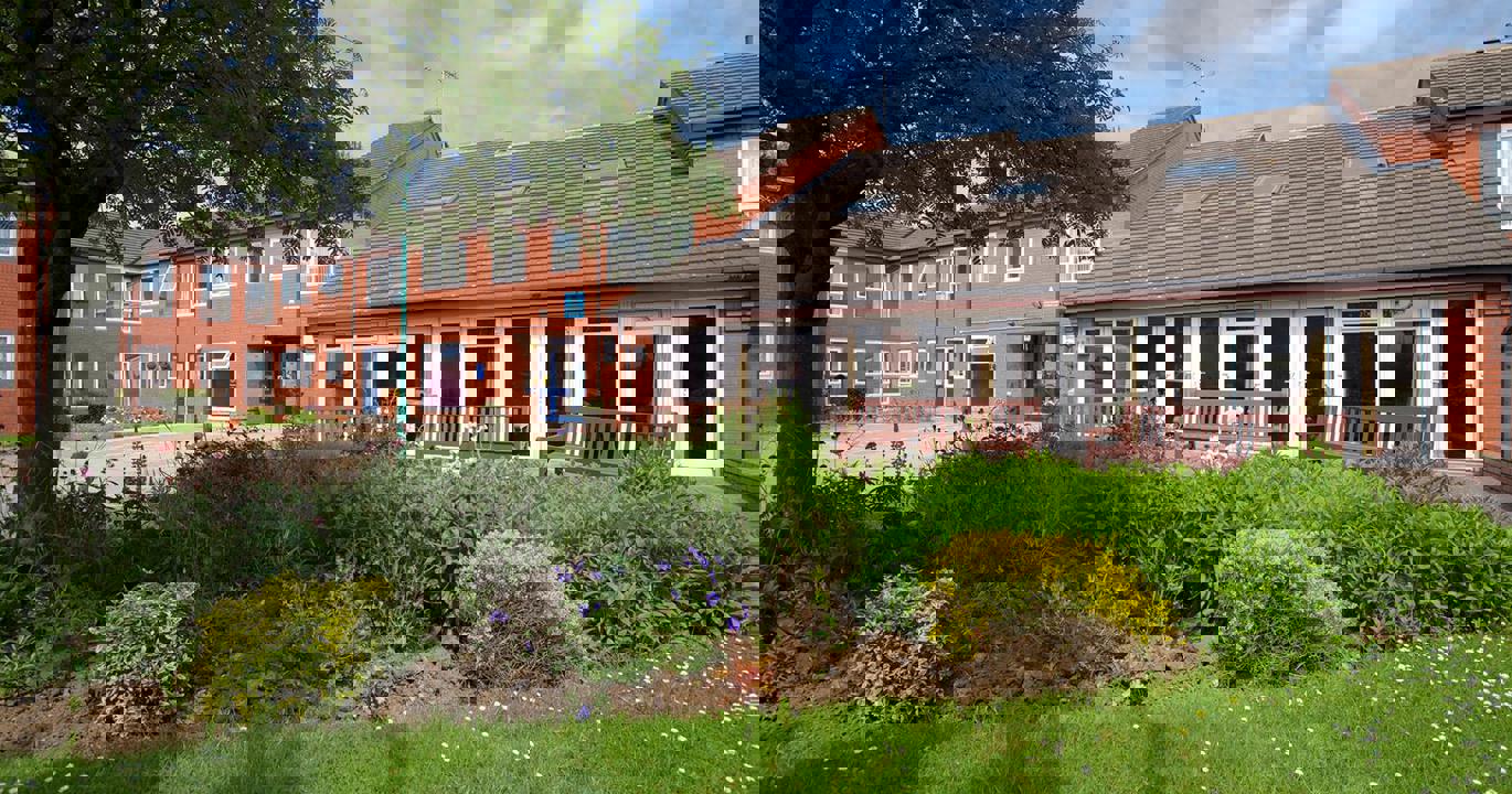 A flowerbed fronted by a lawn and a tree, in the background are three benches and a building with floor-to-ceiling windows and glass doors throughout